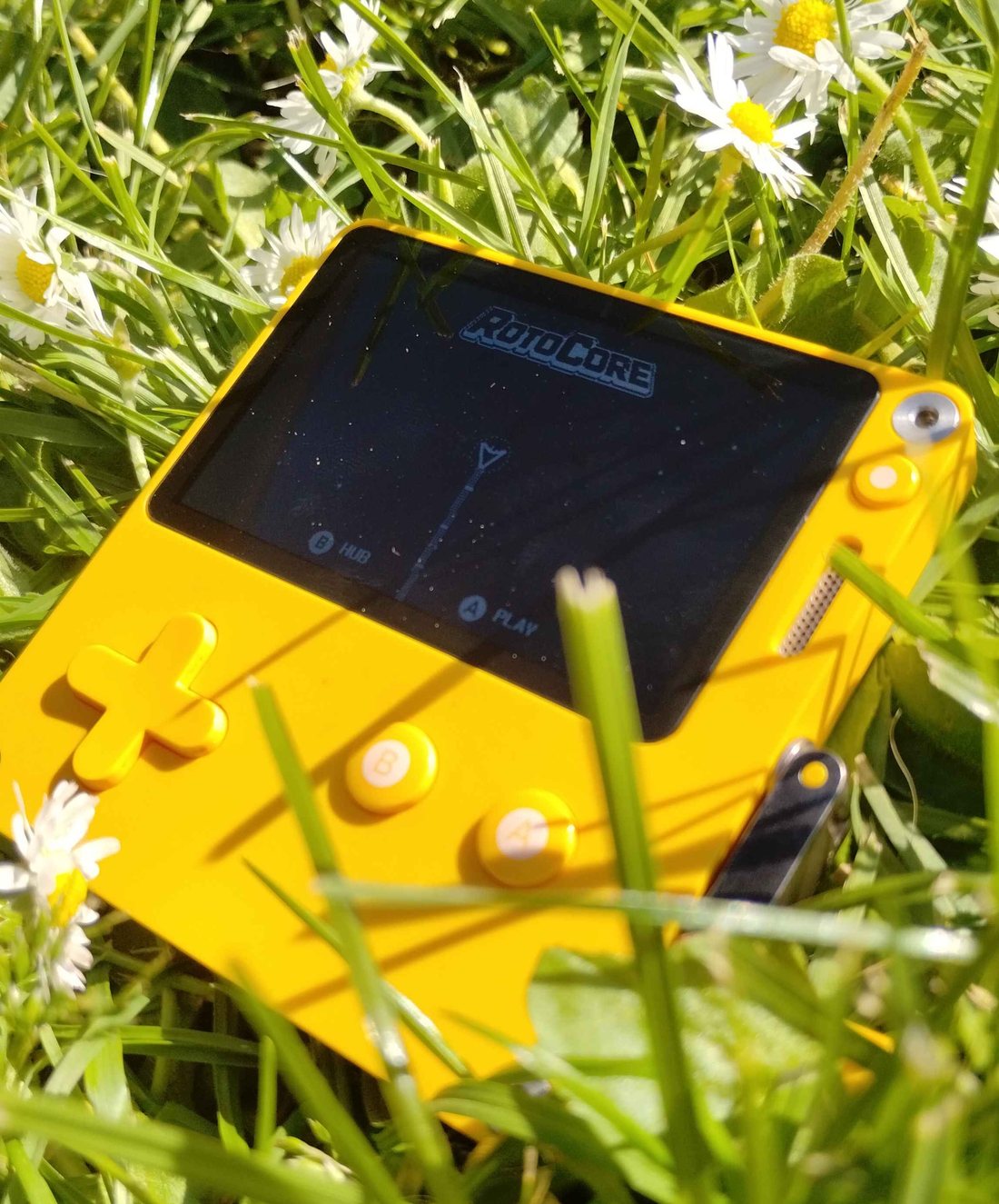 RotoCore running on a yellow Playdate lying in grass among daisies